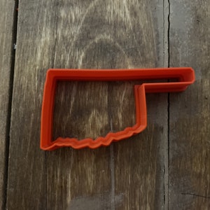 May include: An orange plastic cookie cutter in the shape of a saw. The cutter has a rectangular handle and a jagged blade edge. The cutter is made of a durable plastic material.