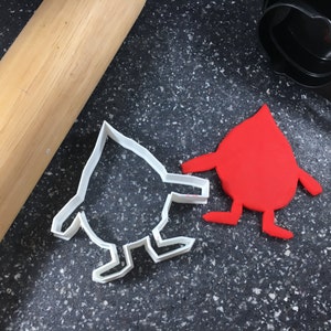 May include: Two cookie cutters, one shaped like a person and the other shaped like a teardrop. The person cookie cutter is white and the teardrop cookie cutter is red.