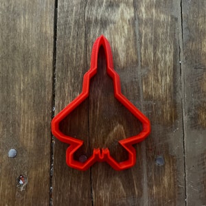Puede incluir: Cortador de galletas de plástico rojo con forma de avión de combate F-22 Raptor. El cortador está sobre una superficie de madera.