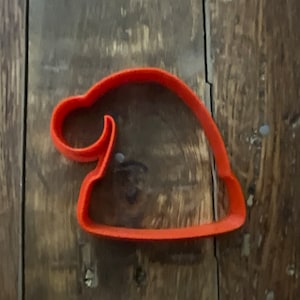 Puede incluir: Cortador de galletas de plástico naranja con forma de gorro de Papá Noel. El cortador tiene un borde curvo y una punta redondeada. Sobre un fondo de madera rústica.