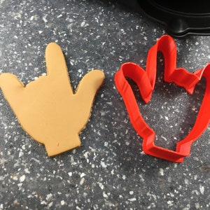 May include: Two cookie cutters, one is red and shaped like a hand giving the 'I love you' sign, the other is tan and shaped like a hand giving the 'rock on' sign.
