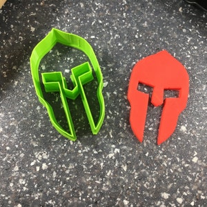 May include: Two plastic cookie cutters shaped like Spartan helmets. One is green and the other is red.