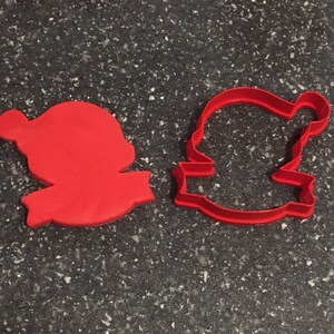 May include: Red plastic cookie cutter shaped like a Santa Claus with a hat and a banner. The cutter is on a dark gray surface.