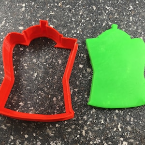 May include: A red plastic cookie cutter shaped like a coffee pot, next to a green coffee pot made of modeling clay.