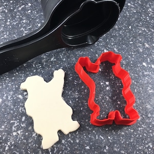Może przedstawiać: Czerwona plastikowa foremka do ciasteczek w kształcie stojącego niedźwiedzia, obok białego ciasteczka wyciętego w tym samym kształcie.