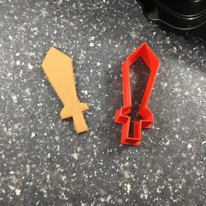May include: Two cookie cutters in the shape of a sword. One is red plastic and the other is yellow plastic.