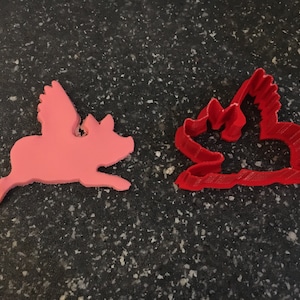 May include: Two cookie cutters in the shape of a flying pig. One is pink and the other is red.