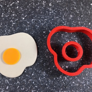 Könnte beinhalten: Ein roter Plastik-Ausstecher in Form eines Spiegeleis, neben einem weißen Plastik-Ausstecher in Form eines Spiegeleis mit einem gelben Eigelb.