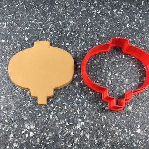 May include: A red plastic cookie cutter shaped like a Chinese lantern, next to a brown plastic cookie cutter of the same shape.