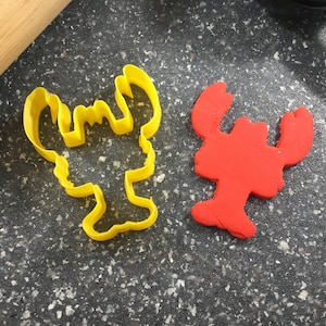 May include: Two lobster cookie cutters, one yellow and one red. The yellow cutter is made of plastic and the red cutter is made of a soft, pliable material.