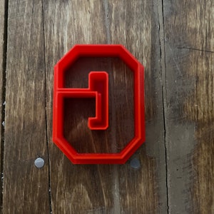 May include: Red plastic cookie cutter shaped like the letter G. The cutter has a slightly raised edge around the outside and a smaller raised edge around the inside of the letter.