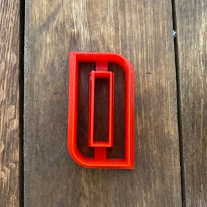 May include: Red plastic cookie cutter shaped like the letter D with a smaller rectangle cut out inside.