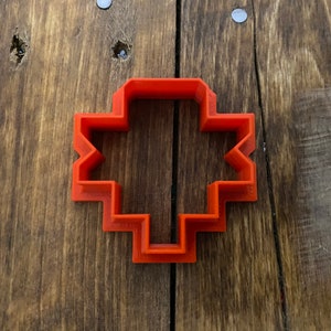 Könnte beinhalten: Ein orangefarbener Plätzchenausstecher aus Kunststoff in Form eines Kreuzes mit geometrischem Muster.