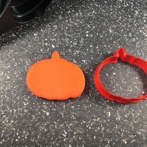 May include: Two pumpkin-shaped cookie cutters, one orange and one red. The orange cutter is solid, while the red cutter is hollow. Both cutters are on a gray speckled surface.