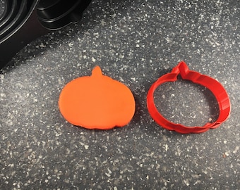 May include: Two pumpkin-shaped cookie cutters, one orange and one red. The orange cutter is solid, while the red cutter is hollow. Both cutters are on a gray speckled surface.