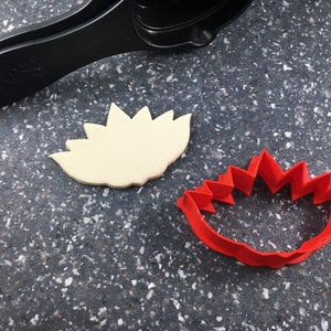 May include: A red plastic cookie cutter shaped like a flower with a white cookie cut out using the cutter.