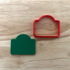 May include: Two cookie cutters on a wooden surface. One is a solid green shape, and the other is a red outline. Both have a rectangular base with a curved top, suitable for making cookies or other treats.