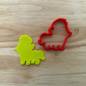 May include: A red plastic lion-shaped cookie cutter and a yellow lion-shaped cookie. The cutter is a simple outline of a lion with a mane, body, and legs. The yellow cookie is a solid shape of the same lion design, perfect for baking.