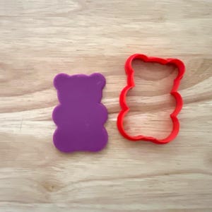 May include: A purple gummy bear-shaped cookie cutter and a red outline of the same shape sit on a light wood surface. The purple cutter is solid, while the red one is an open outline.