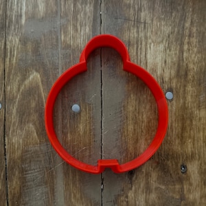 May include: A bright red cookie cutter in the shape of a Christmas ornament. The cutter is made of plastic and has a rounded top and a small rectangular opening at the bottom of the circle. The cutter is on a wooden surface.
