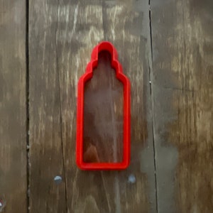 Puede incluir: Cortador de galletas de plástico rojo con forma de botella, con la parte superior redondeada y el cuerpo rectangular.