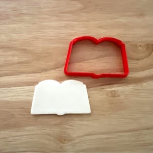 May include: A red plastic cookie cutter in the shape of an open book sits next to a white fondant book. The items are on a light brown wooden surface. The cookie cutter is a simple outline, while the fondant book has a detailed shape.