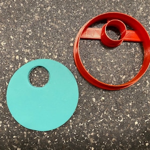 Pode incluir: Dois cortadores de biscoitos, um vermelho e outro azul turquesa, ambos com formato circular e um círculo menor cortado no centro.