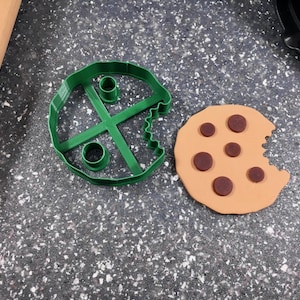 May include: Green plastic cookie cutter shaped like a cookie with a bite taken out of it. Next to it is a brown cookie with chocolate chips and a bite taken out of it.