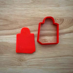 May include: Two red cookie cutters in the shape of a shopping bag, one solid and one outline, sit on a wooden surface. The cutters are made of a flexible, food-safe plastic, and are designed for baking.