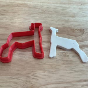 May include: A red plastic cookie cutter in the shape of a dog, next to a white dog-shaped cookie. The cookie cutter is a simple outline, while the cookie has more detail. Both are on a wooden surface.