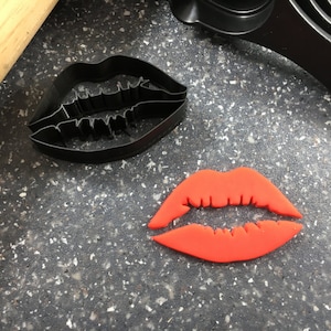 Peut inclure: Une découpe de lèvres en fondant rouge, à côté d'un emporte-pièce en métal noir en forme de lèvres.