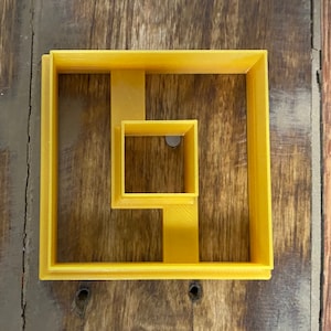 May include: A yellow plastic cookie cutter shaped like a square with a smaller square cut out of the center.