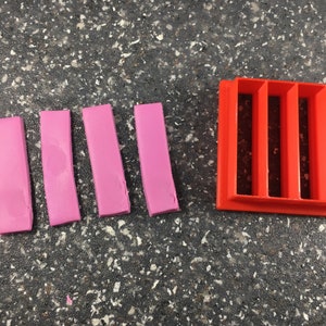 May include: Five pink rectangular pieces of clay next to a red plastic mold with five rectangular slots.