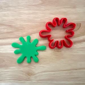 May include: Two splash-shaped cookie cutters. One is bright green and solid, the other is red and outlines the same shape. The cutters are made of plastic and sit on a light brown wooden surface.