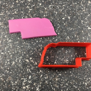May include: A red plastic cookie cutter in the shape of the state of Nebraska. A pink plastic outline of the state of Nebraska is also in the image.