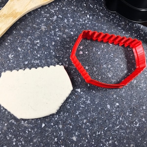 Peut inclure: Un emporte-pièce en plastique rouge en forme de sous-vêtements et une pâte à biscuits blanche sur une surface grise mouchetée.