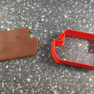 Könnte beinhalten: Ein roter Plastik-Ausstecher in Form eines Bootes, neben einem braunen Keksteig-Ausschnitt in der gleichen Form.