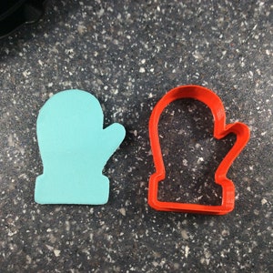 May include: A light blue mitten-shaped object and a red mitten-shaped cookie cutter are displayed on a speckled gray surface. The objects are side-by-side, showcasing the shape and design for baking or crafting.