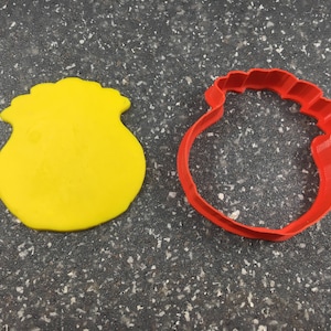 May include: Two cookie cutters, one yellow and one red. The yellow cutter is shaped like a pouch or bag. The red cutter is shaped like a rounded, open-top container.