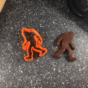 May include: Two cookie cutters shaped like Bigfoot, one orange and one brown, on a dark gray speckled surface.