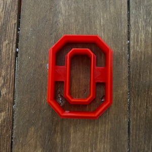 May include: Red plastic cookie cutter shaped like the letter "O" with a smaller "O" inside. The cutter is on a wooden surface.