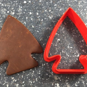 May include: A red plastic cookie cutter shaped like an arrowhead, next to a brown cookie cut with the same shape.