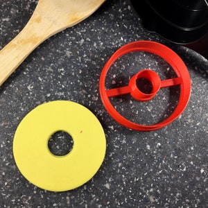 May include: A yellow donut-shaped object and a red circular cookie cutter with a central circle and a connecting bar are on a speckled gray surface. A wooden spoon and black measuring cup are also visible.