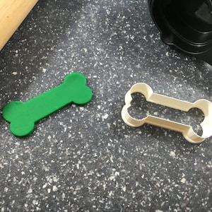 May include: A green plastic bone-shaped cookie cutter and a white plastic bone-shaped cookie cutter on a gray speckled surface.