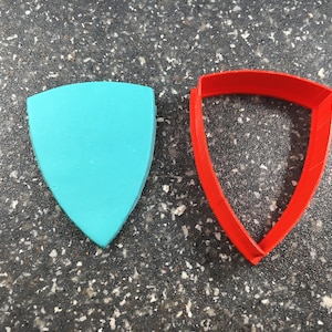 May include: A red plastic cookie cutter shaped like a shield, next to a teal blue plastic shield.