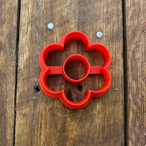Puede incluir: Un cortador de galletas de plástico rojo en forma de flor con un agujero redondo en el centro.