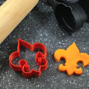 Könnte beinhalten: Zwei Ausstecher in Form einer Fleur-de-lis. Einer ist aus rotem Kunststoff und der andere aus orangefarbenem Fondant.