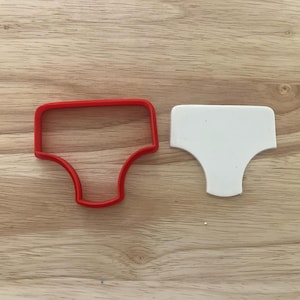 Peut inclure: Un emporte-pièce en plastique rouge en forme de sous-vêtement est posé à côté d'une découpe de biscuit blanche de la même forme. Les objets sont sur une surface en bois brun clair.