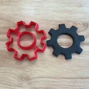 May include: A red plastic gear-shaped cookie cutter and a black gear-shaped object are displayed on a wooden surface. The cookie cutter has a circular center and a toothed outer edge. The black object has a similar design.