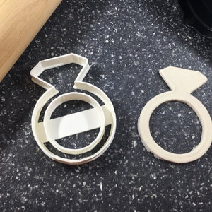 May include: Two white plastic cookie cutters shaped like engagement rings. The cutter on the left has a detailed diamond design, while the cutter on the right is a simple ring shape.